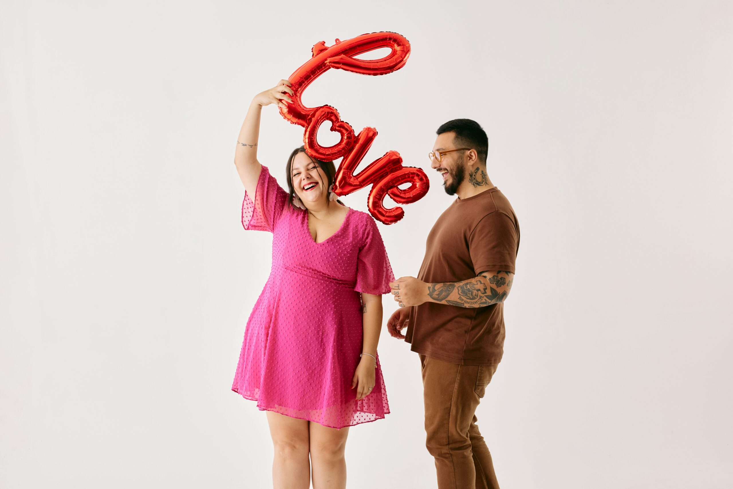 A joyful couple shares a laugh while holding a red balloon spelling "Love." The woman wears a vibrant pink dress, and the man sports a brown T-shirt with tattoos, creating a fun and lighthearted Valentine's Day vibe.